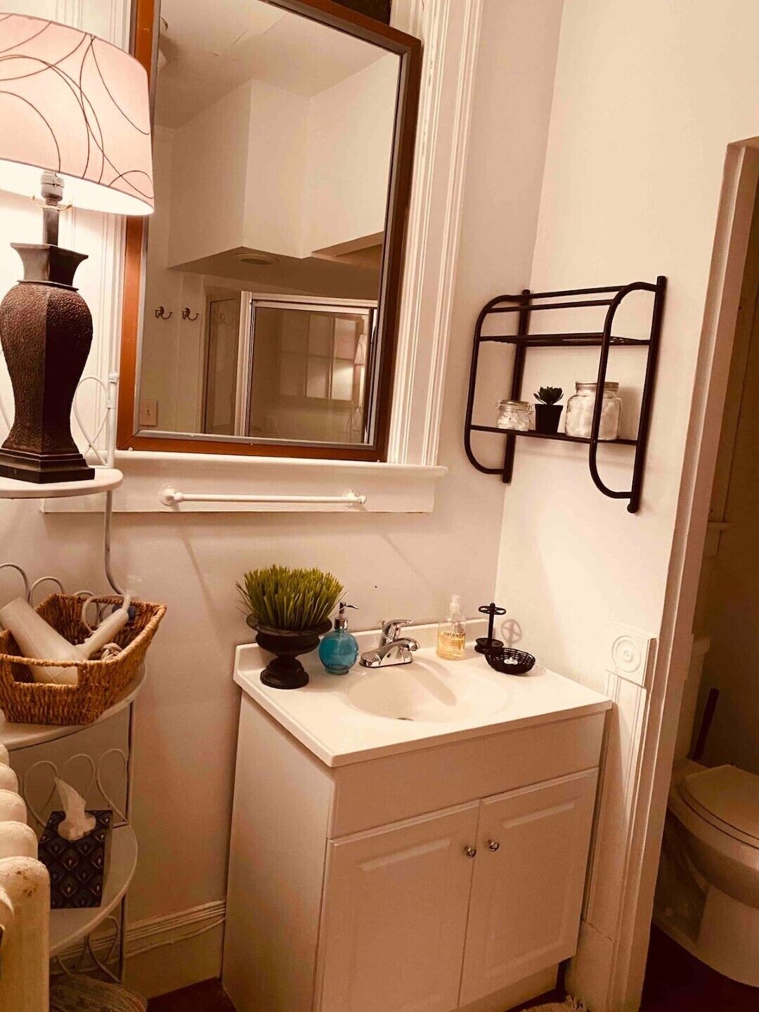 Un bagno con un lavandino bianco, uno specchio, uno scaffale con articoli da toilette e una lampada.