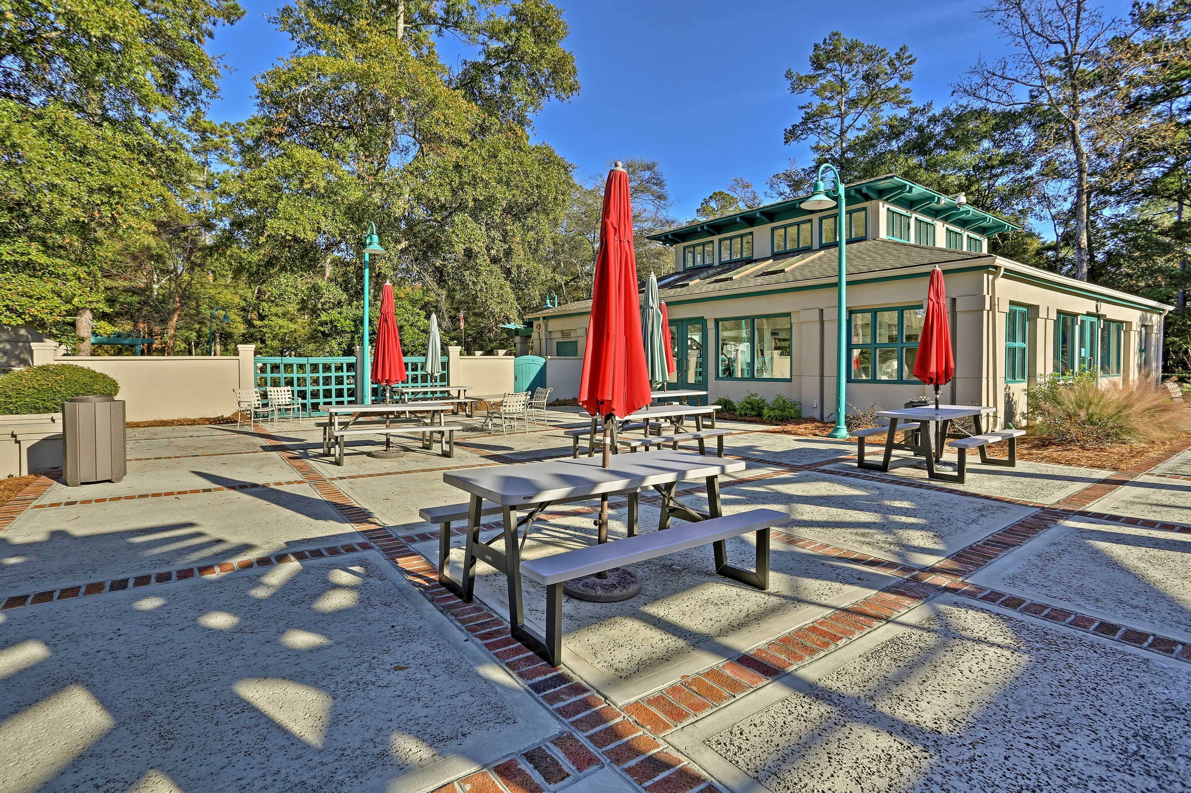 An outdoor patio with picnic tables and red umbrellas, surrounded by trees and a building with green trim.