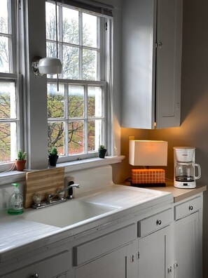 A kitchen with a white countertop, a sink, a coffee maker, and a window with a view of trees. - Pretty Little Apt by the Train II (Winchester)