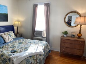 A bedroom with a bed, a dresser, a mirror, a window with curtains, a radiator, and a lamp.