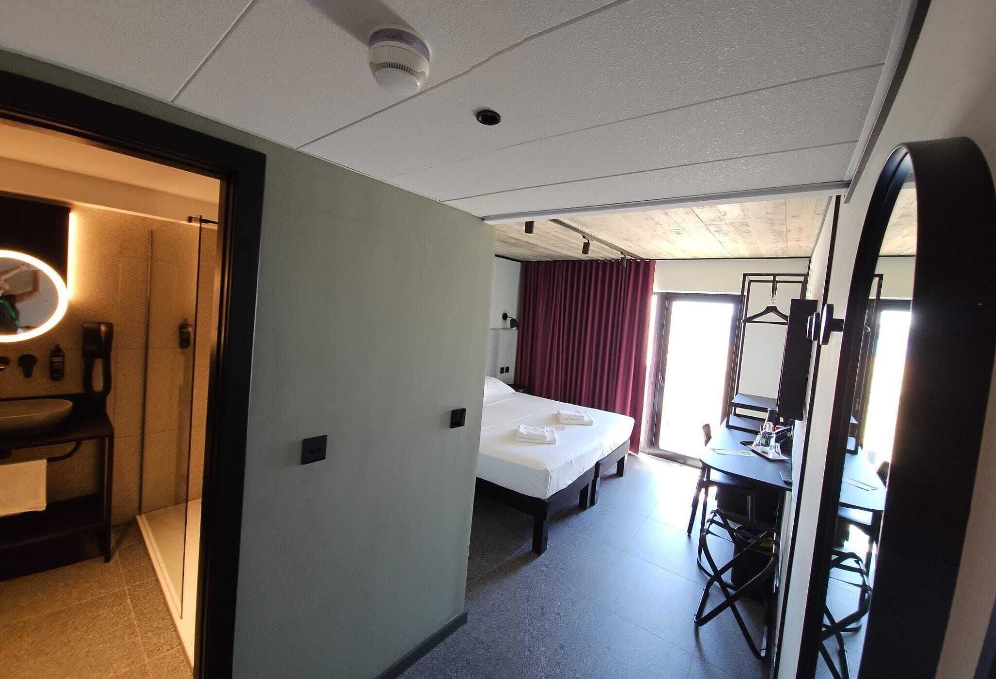 Une chambre d’hôtel comprenant un lit, un bureau avec une chaise, un miroir et une salle de bain avec un lavabo et un luminaire rond.