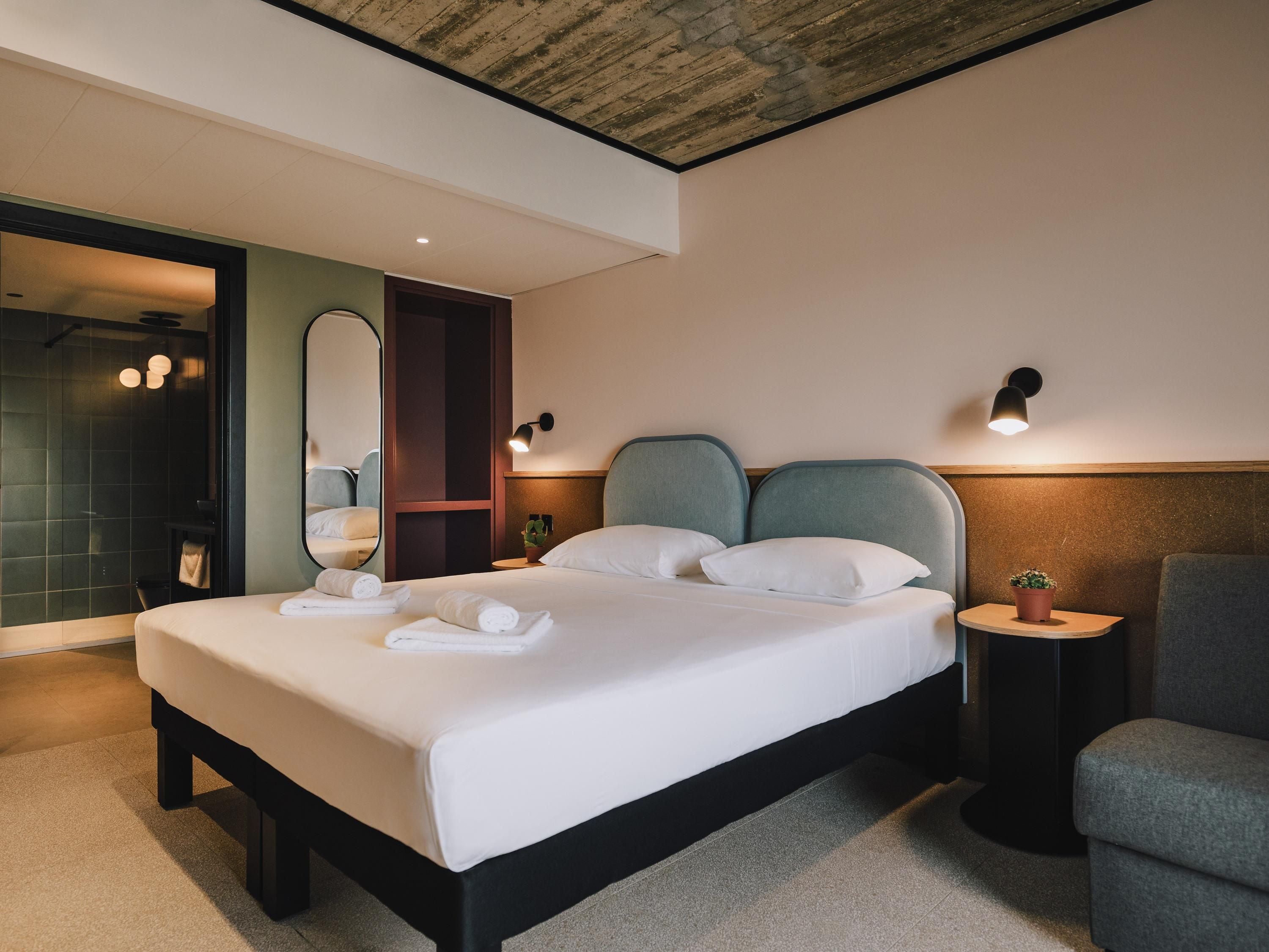 Una habitación de hotel moderna con una cama grande, una mesita de noche con una planta, una silla y un box de ducha.