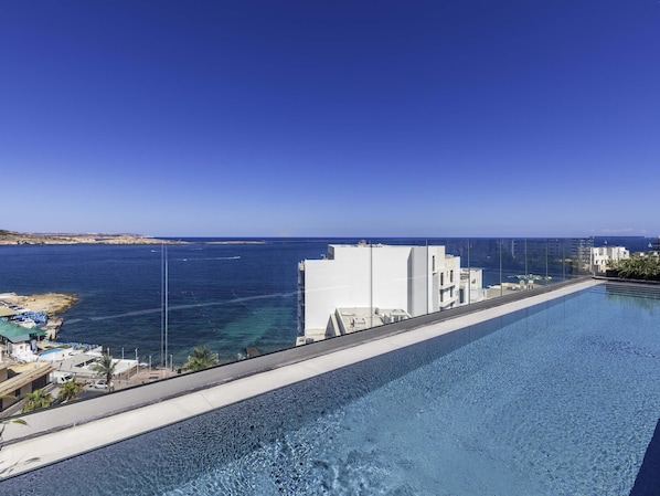 Una piscina infinita en la azotea con vistas despejadas al mar y un edificio blanco al fondo.