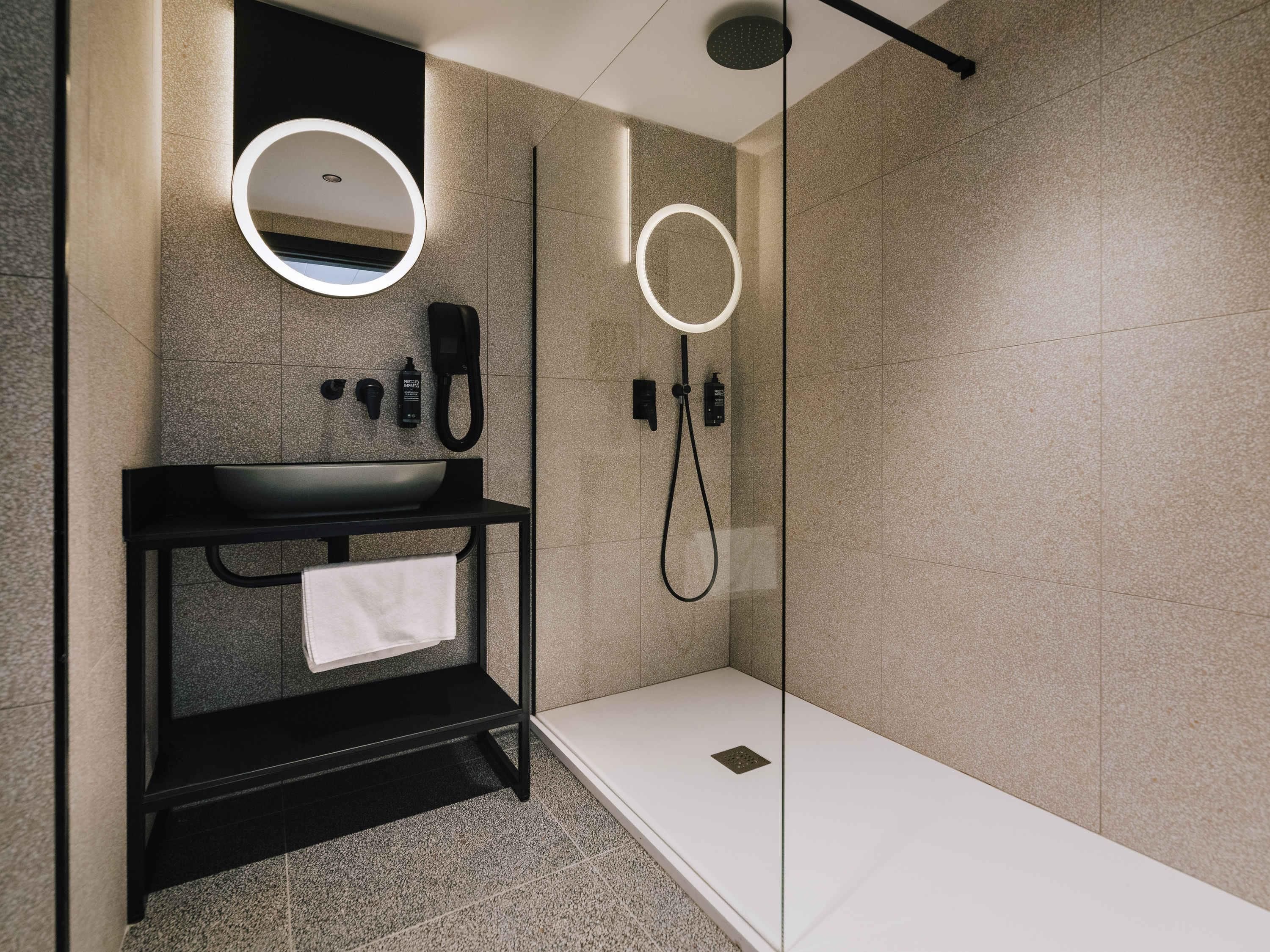 A modern bathroom with a black vanity, a mirror with a circular light, a handheld showerhead, and a glass-enclosed shower area.