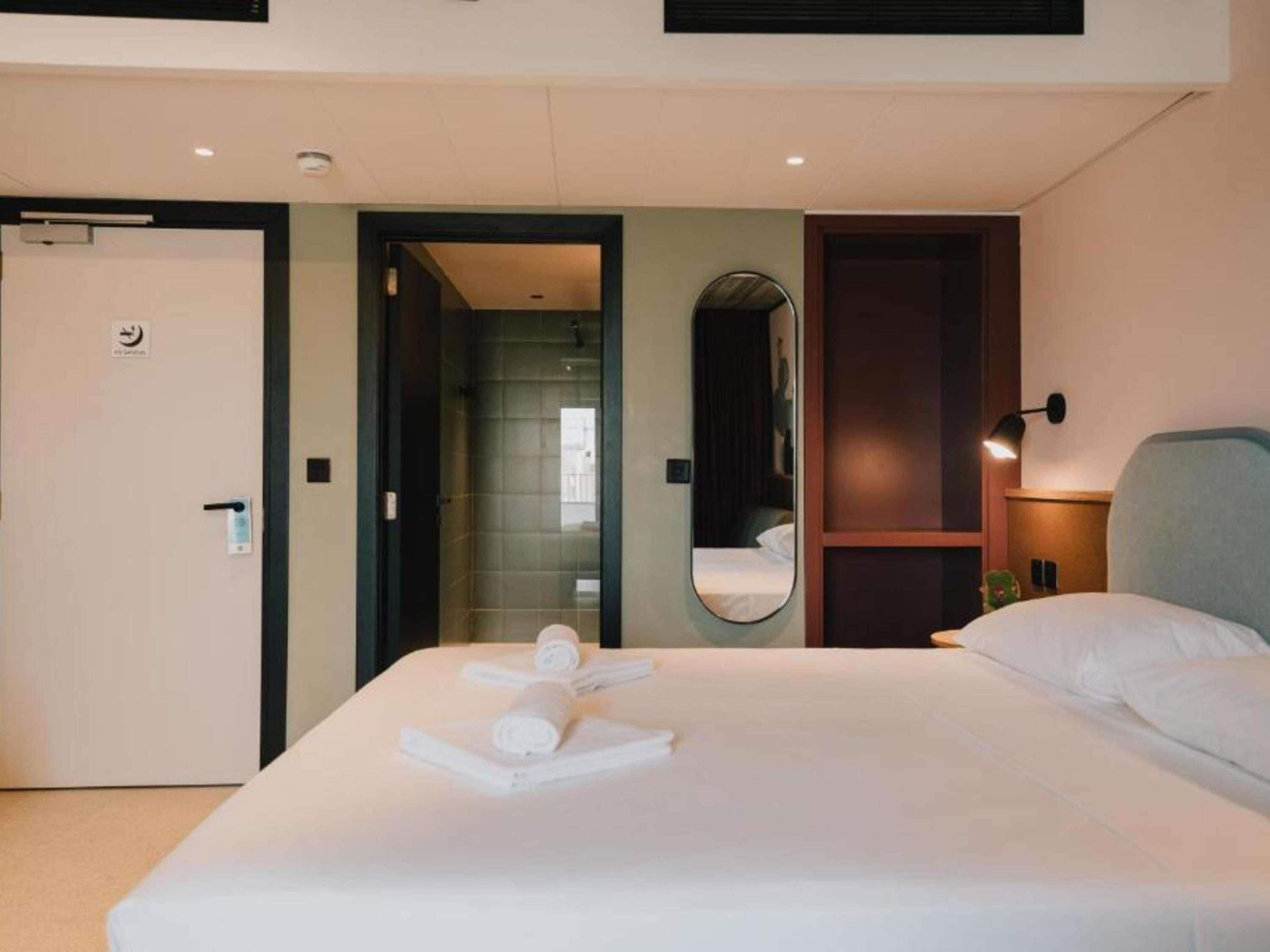 A hotel room with a bed, a mirror, a bedside table, and a door.