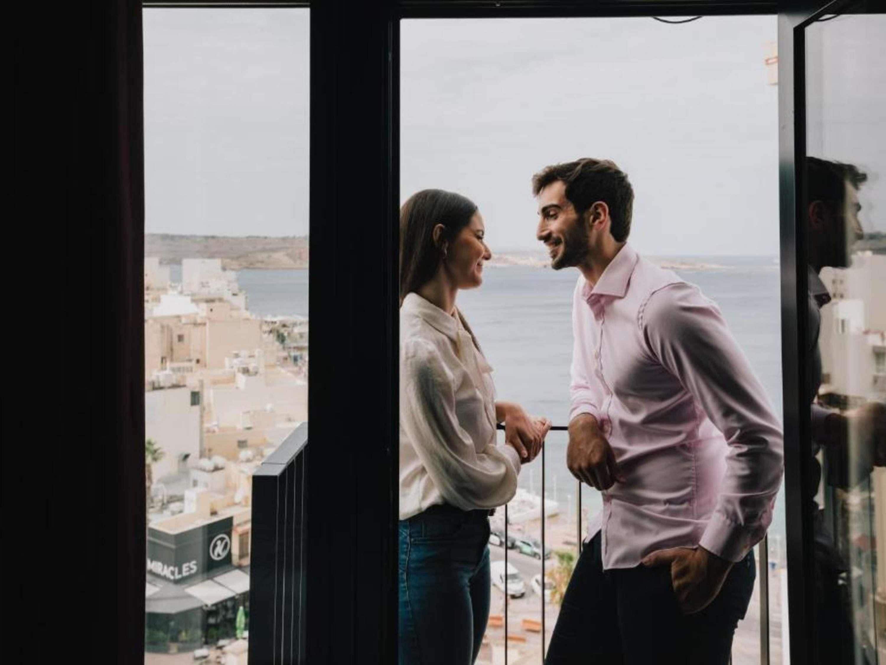 A couple standing by a window overlooking a cityscape with buildings and a body of water.