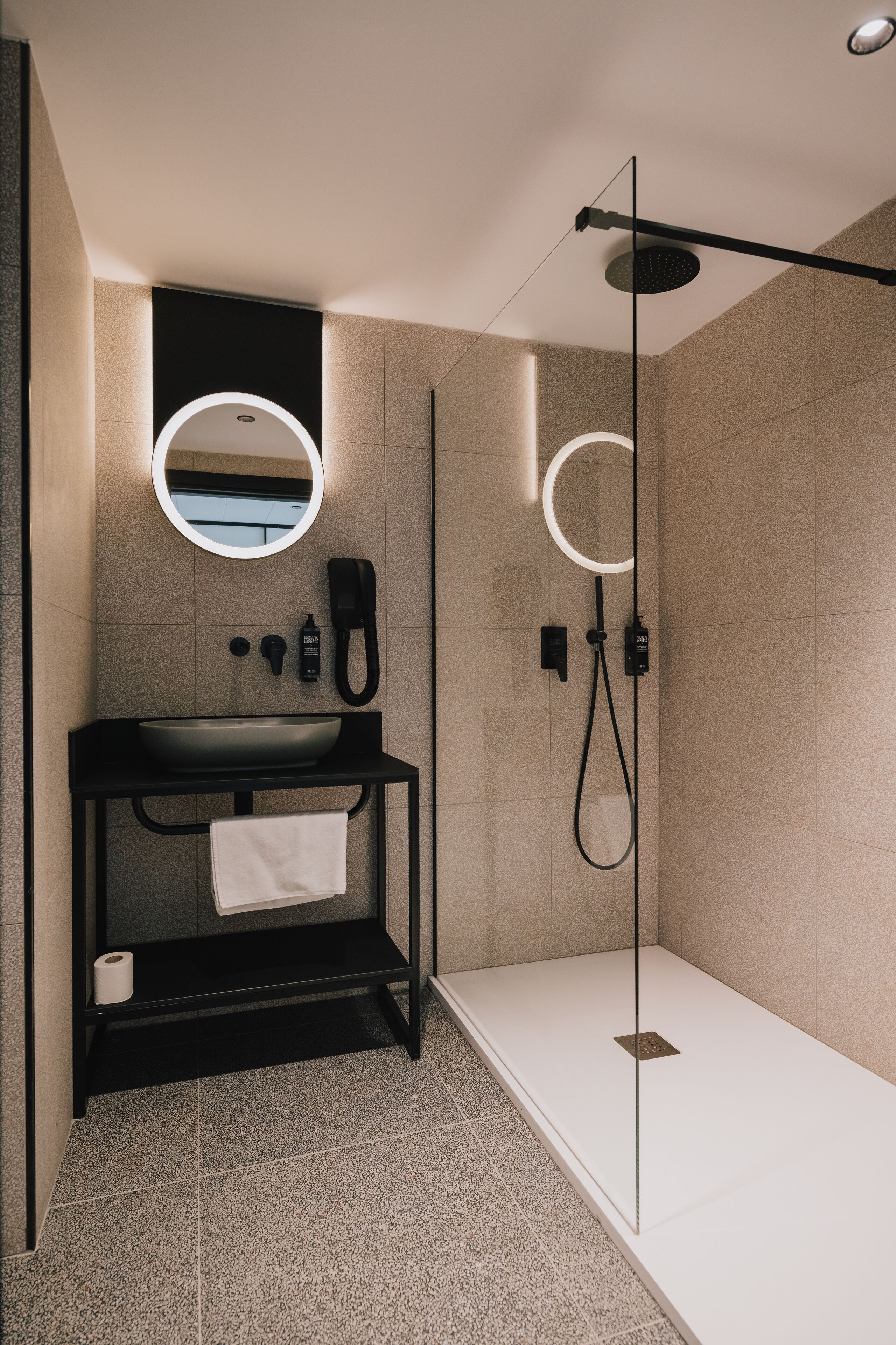 Een moderne badkamer met een zwart badkamermeubel, een ronde spiegel, een handdouche en een douche met glazen wanden.