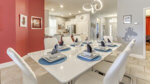 A dining area with a long table set for six, white chairs, and a modern chandelier. - Marvel Villa - 20 minutes to Disney (Davenport)