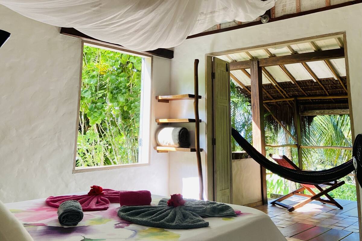 Una habitación con cama, una hamaca, un ventanal con vistas a follaje y un estante de madera.