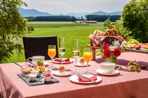 Une table de pique-nique dressée avec une variété de plats et de boissons, entourée d’un paysage pittoresque composé d’un champ verdoyant et de montagnes.