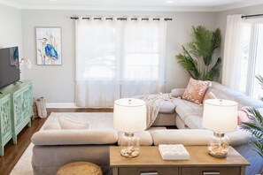 Uma sala de estar moderna com um sofá seccional, duas luminárias sobre uma mesinha de apoio, uma televisão num armário e uma grande janela com cortinas.