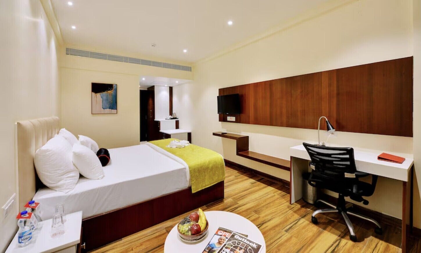 Une chambre d’hôtel avec un lit, un bureau, une chaise, une télévision et une petite table sur laquelle se trouvent un bol de fruits et un magazine.