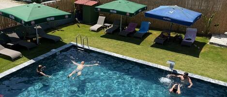 A swimming pool with clear water, surrounded by lounge chairs and umbrellas, set against a backdrop of residential buildings and a mountain.