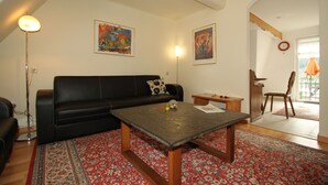 Ein Wohnzimmer mit einem schwarzen Ledersofa, einem Couchtisch aus Holz und einem roten Teppich mit Muster. An den Wänden hängen zwei Gemälde, eine Stehlampe und ein kleiner Tisch mit einem Stuhl.
