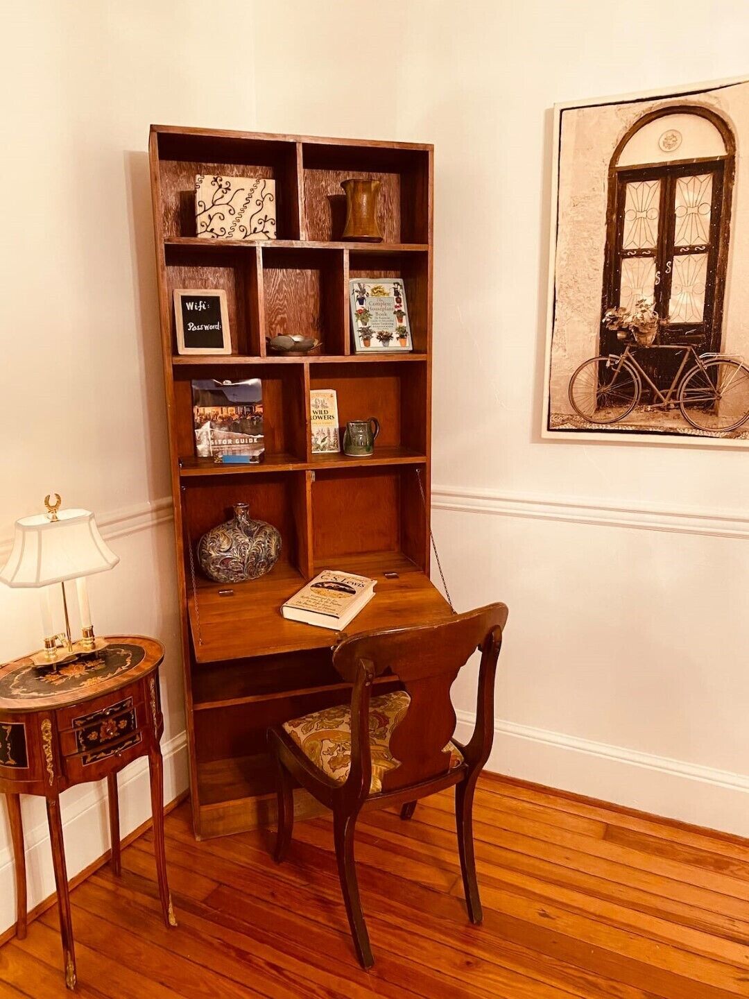 A wooden bookshelf with various books and decorative items, a wooden chair, a small table with a lamp, and a framed picture of a bicycle on the wall.