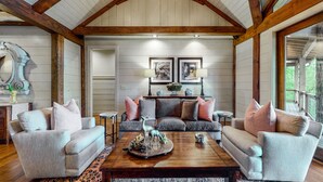 A spacious living room with a high wooden ceiling, a grey sofa set, a wooden coffee table with decorative items, and framed pictures on the wall. - Looks Like Love! on Valentine Lane: 2 Levels of Luxury Home w Mtn Views, Porches (Highlands)