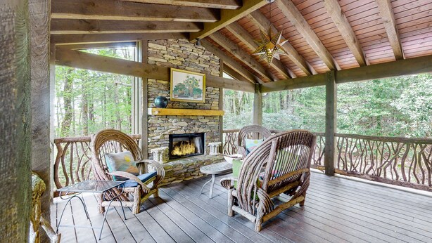 A covered porch with wooden beams, a stone fireplace, wicker furniture, and a view of the forest. - Looks Like Love! on Valentine Lane: 2 Levels of Luxury Home w Mtn Views, Porches (Highlands)