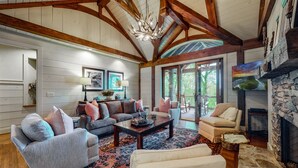 A spacious living room with a high ceiling, wooden beams, a fireplace, and a large window overlooking a deck. - Looks Like Love! on Valentine Lane: 2 Levels of Luxury Home w Mtn Views, Porches (Highlands)