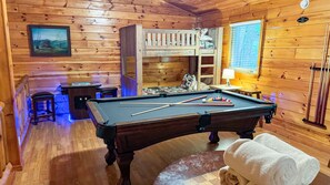 Ein Billardtisch mit Spielstöcken und Kugeln, ein Etagenbett, ein gerahmtes Bild und ein Fenster mit Jalousien.