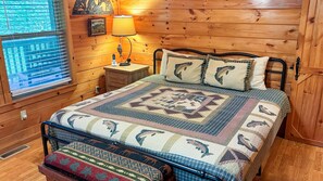 Ein Schlafzimmer einer Holzhütte mit einem Bett, einem Nachttisch, einer Lampe und einem Fenster mit Jalousien.