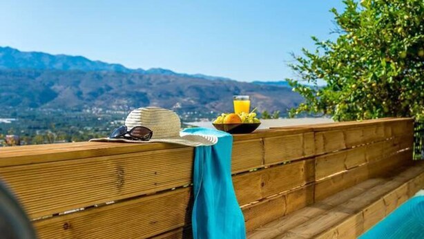 Um deck de madeira com piscina, uma mesa com chapéu e óculos de sol, e uma tigela de frutas.