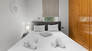Quarto com cama, duas almofadas, mesa de cabeceira com abajur, janela com persiana e guarda-roupa de madeira.