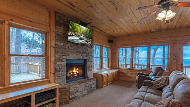 Ein gemütliches Blockhauss-Interieur mit Kamin aus Stein, Holzmöbeln und großen Fenstern mit Blick ins Freie.