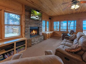 A cozy cabin interior with a stone fireplace, wooden furniture, and large windows offering views of the outdoors. - A Bear`s Lair - Mountain View - Blue Ridge, GA (Mineral Bluff)
