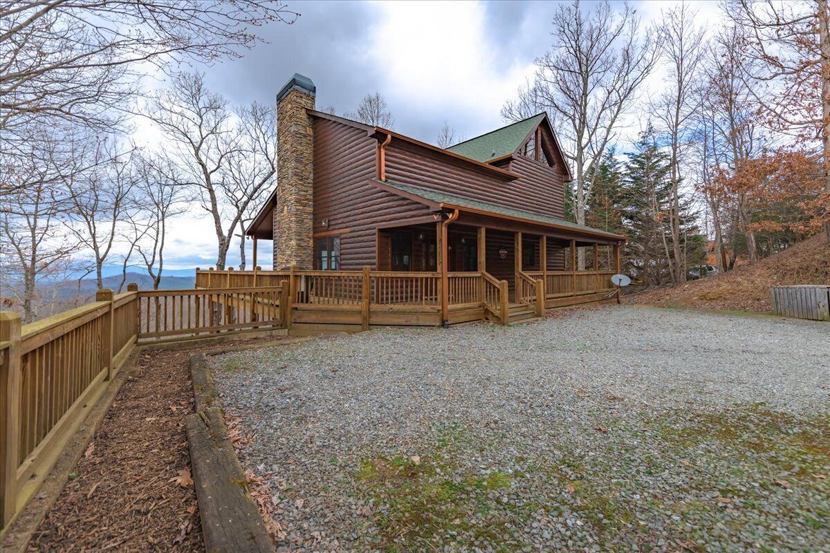 A log cabin with a stone chimney, wooden deck, and a gravel driveway.