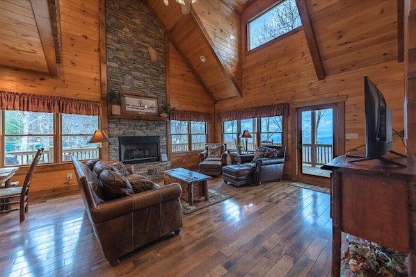 A spacious log cabin interior with a fireplace, leather furniture, and large windows offering views of the outdoors.