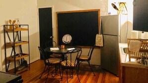 A compact kitchen with a dining table, chairs, a clock, a refrigerator, and a shelf with kitchen utensils.