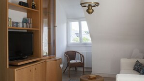 A living room with a wooden entertainment unit, a television, a wicker ottoman, a white sofa, and a ceiling light fixture. - Self-contained apartment in Breton house (Loctudy)
