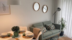 A dining area with a round table set for four, surrounded by white chairs. A grey sofa with patterned cushions is against a wall with two round mirrors. A potted plant is in the corner, and a floor lamp is near the window with sheer curtains. - Oporto City Center Apartment with terrace (Porto)