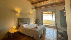 Une chambre avec un grand lit, des poutres apparentes en bois au plafond, une table de chevet avec une lampe, et une vue sur l’extérieur grâce à une porte-fenêtre coulissante.