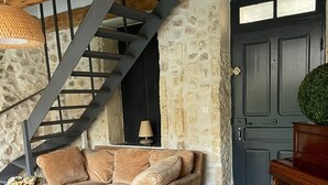 Un salon chaleureux avec un mur en pierre, un escalier en bois, un canapé avec des coussins, une table basse et un piano.