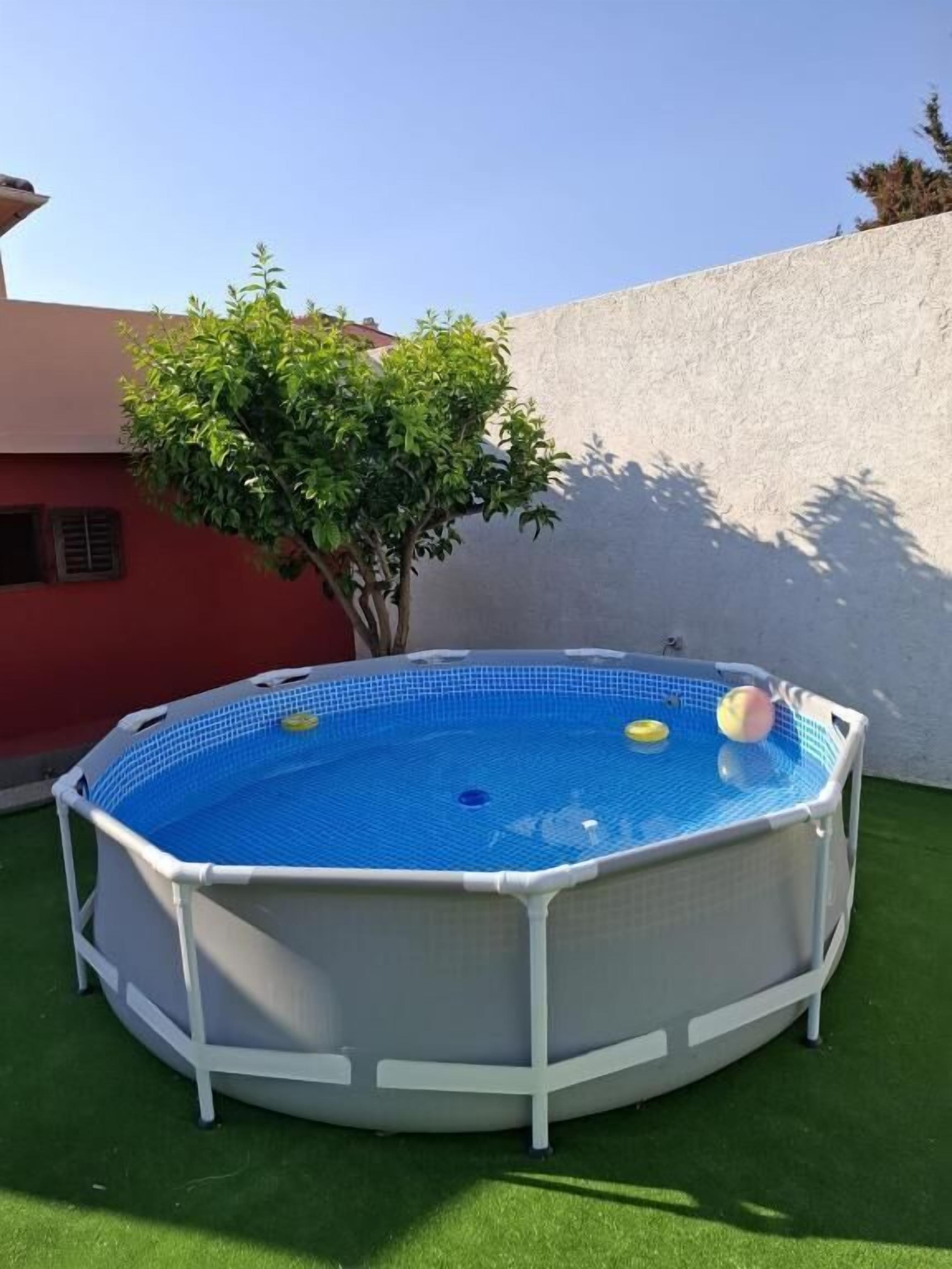 Piscina inflável redonda, com interior azul e estrutura branca, rodeada por grama sintética e uma árvore.