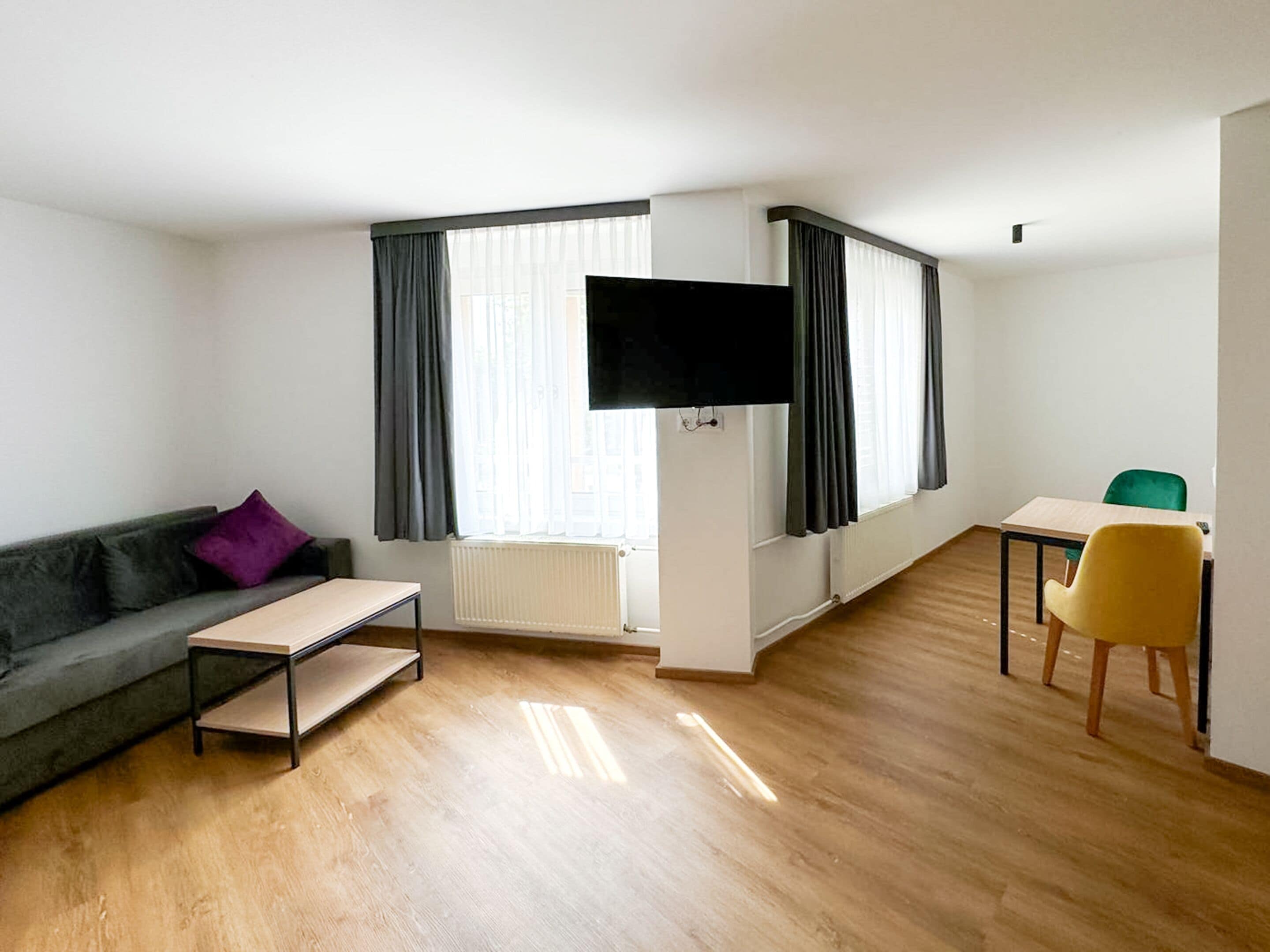 A modern living room with a sofa, a small table, a TV mounted on the wall, and a dining area with a table and chairs.