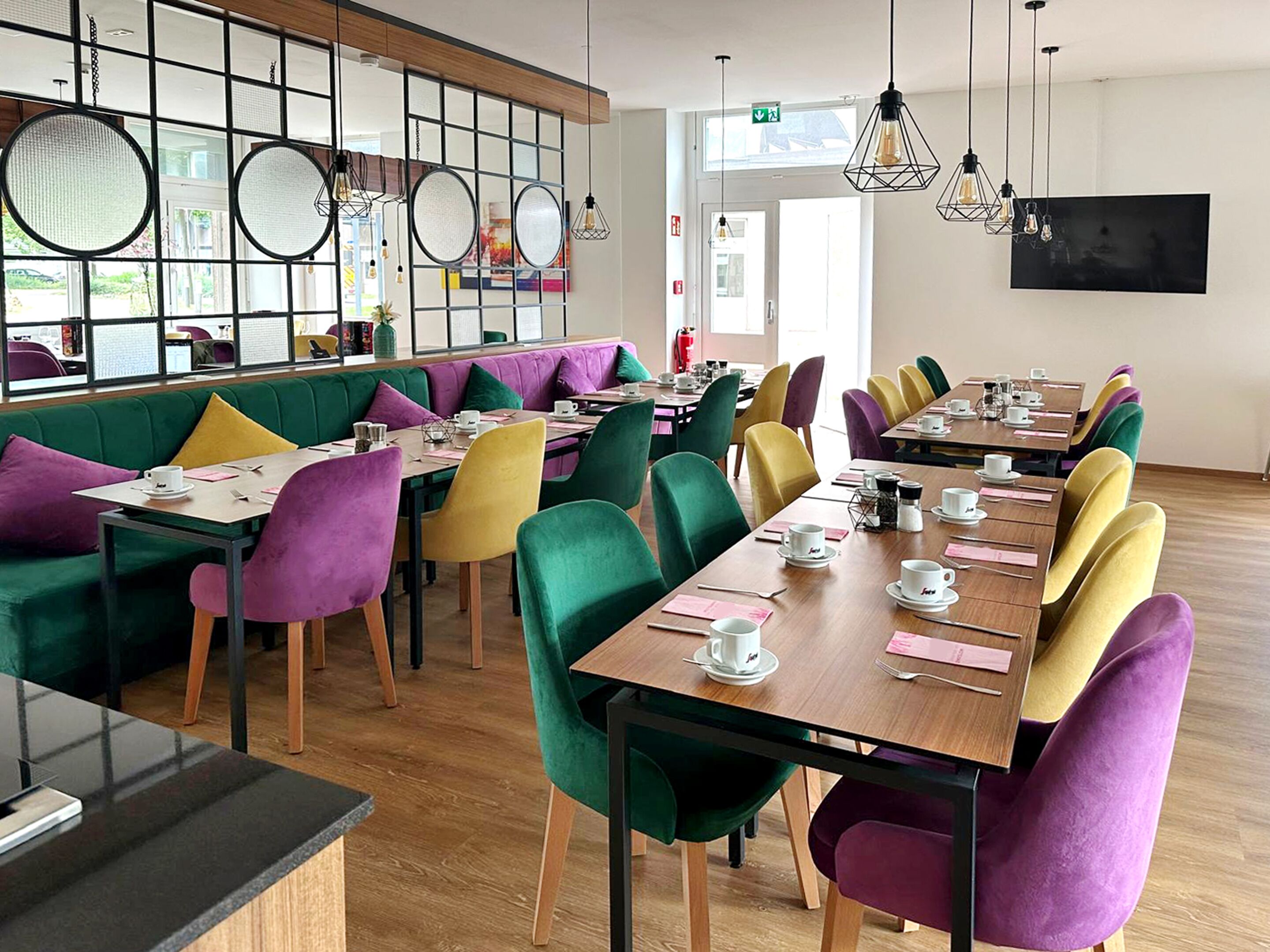Un espace repas moderne avec des sièges aux couleurs vives et de longues tables, comprenant un mélange de tables rondes et rectangulaires, ainsi qu’une variété de chaises éclatantes.