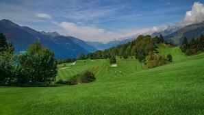 A panoramic view of a lush green golf course with rolling hills, distant mountains, and a clear sky. - Hotel mischun (Brigels)