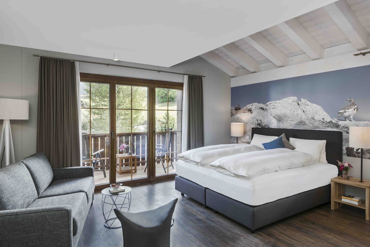 A modern hotel room with a large bed, a sofa, a small table, and a balcony with a view of greenery.