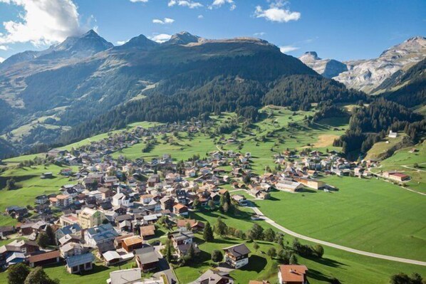 Une petite ville nichée dans une vallée, avec des maisons et des bâtiments dispersés dans de vastes champs verdoyants, le tout entouré de montagnes.