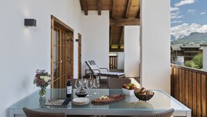 A glass-top table with a bottle of wine, wine glasses, plates, cutlery, a bowl of fruit, and a vase of flowers on a balcony with wooden railings, a wicker chair, and a view of mountains. - Hotel mischun (Brigels)