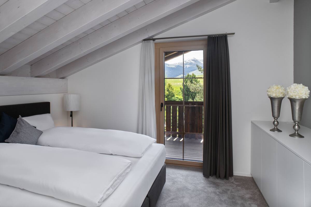 A bedroom with a large bed, a nightstand with a lamp, a dresser, and a view of a balcony with a mountain backdrop.
