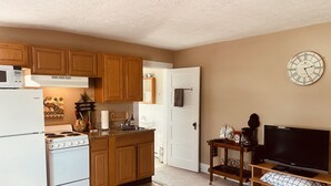 Una cocina compacta con armarios de madera, un refrigerador blanco y una estufa. Hay una cafetera en la encimera y un televisor sobre un soporte de madera.