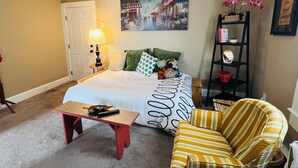 Un dormitorio con una cama, una mesita de noche, una lámpara, una silla, una estantería y un cuadro en la pared.