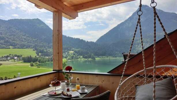 A wooden balcony with a hanging chair, a table with breakfast items, and a view of a lake and mountains. - Aparthotel Buchauer.Tirol in a natural paradise (Thiersee)