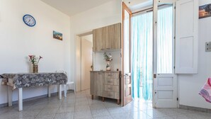 A room with a tiled floor, a dining table with a floral tablecloth, a wooden cabinet, a clock, and a door with curtains.