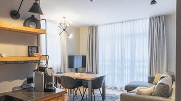 Habitación de hotel moderna con una mesa de comedor, sillas, un sofá, una máquina de café, un televisor y ventanas grandes con cortinas.