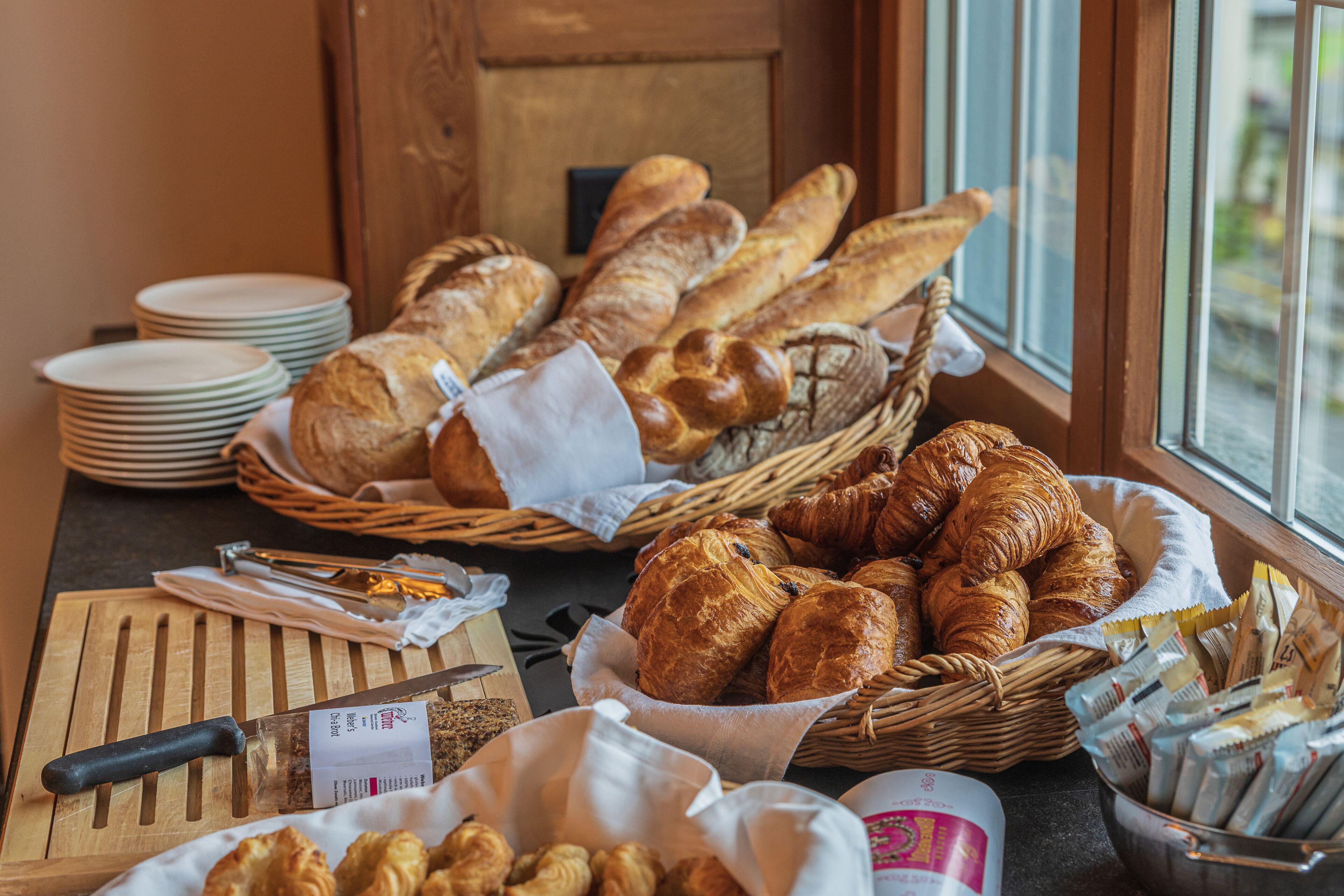 Un bufé de desayuno con panes y pasteles diversos, una tabla de cortar con cuchillo y una cesta de periódicos.