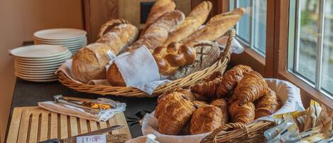 Un bufé de desayuno con panes y pasteles diversos, una tabla de cortar con cuchillo y una cesta de periódicos.